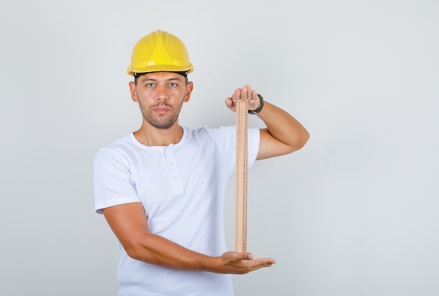 Male builder in white t-shirt, security helmet holding wooden ruler, front view.