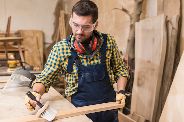 Free photo a male carpenter cleaning the wooden plank with brush in the workshop