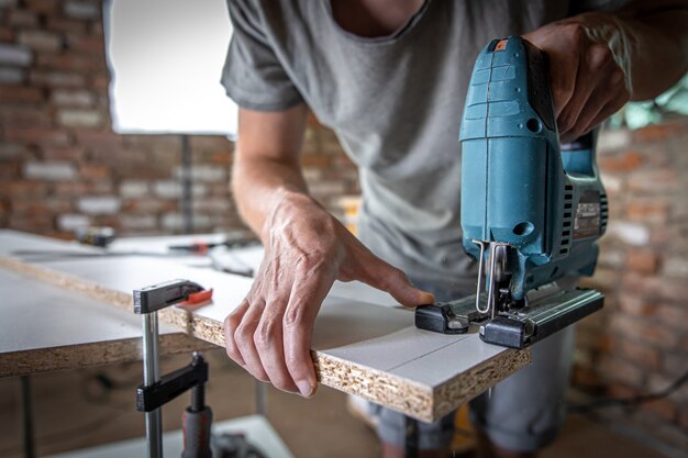 A male carpenter cuts a wood with an electric jigsaw, working with a tree.