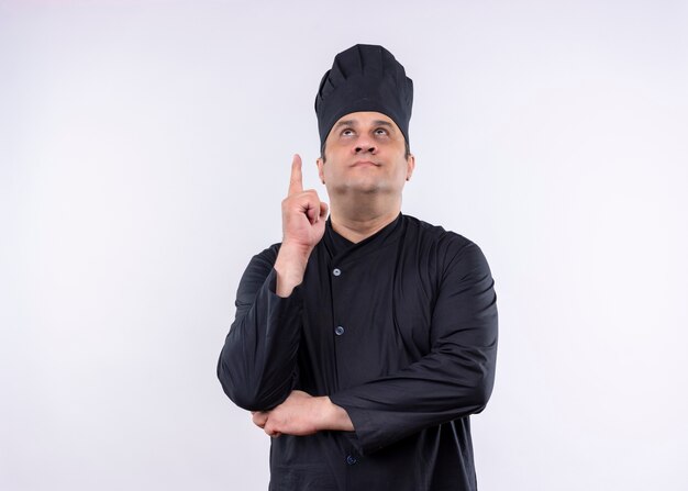 Male chef cook wearing black uniform and cook hat pointing with index finger up looking up standing over white background