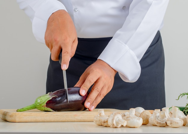 Male chef cutting eggplant on wooden board in uniform and apron in kitchen