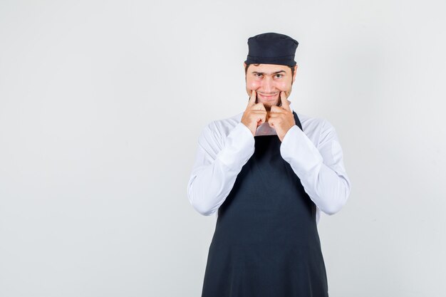 Male chef forcing a smile on face in uniform, apron and looking gloomy. front view.