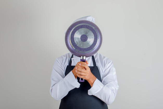 Free photo male chef hiding behind frying pan in hat, apron and uniform