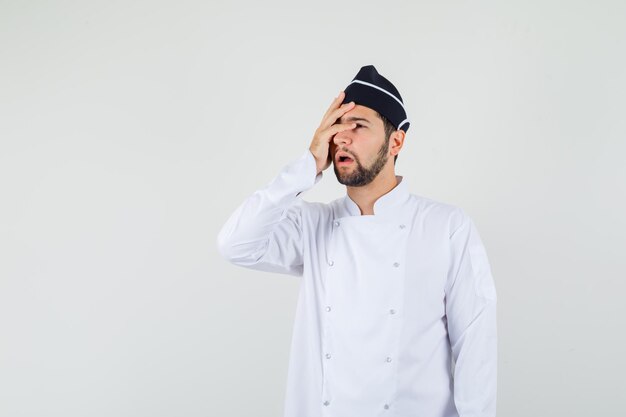 Male chef holding hand on his face in white uniform and looking worried , front view.