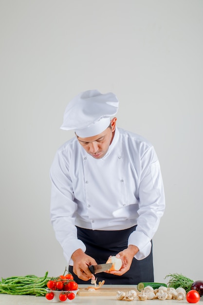 Male chef peeling onion on wooden board in uniform, apron and hat in kitchen