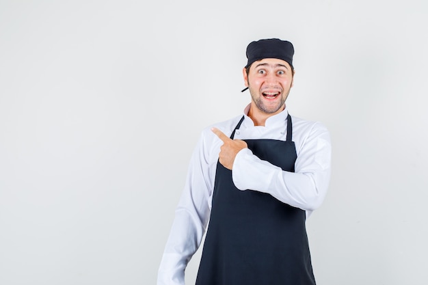 Male chef pointing back in uniform, apron and looking cheerful. front view.