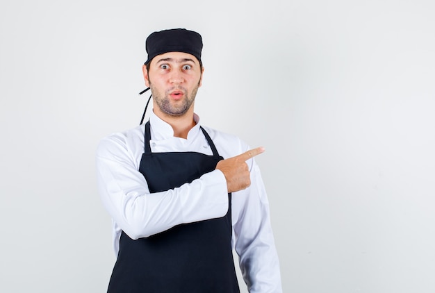 Male chef pointing finger to side in uniform, apron and looking surprised. front view.