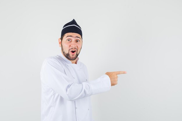 Male chef pointing to the side in white uniform and looking amazed. front view.