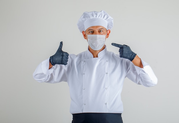 Male chef in uniform, apron and hat showing mask and thumbs up and looking careful