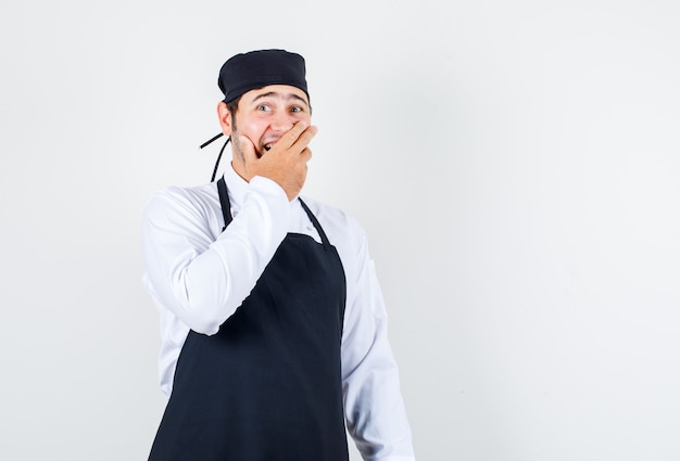 Male chef in uniform, apron holding hand on mouth and looking surprised , front view.