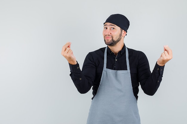 Male cook gesturing with fingers in shirt, apron and looking cheerful , front view.