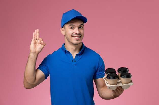 male courier in blue uniform holding coffee cups and posing on pink, worker uniform service delivery