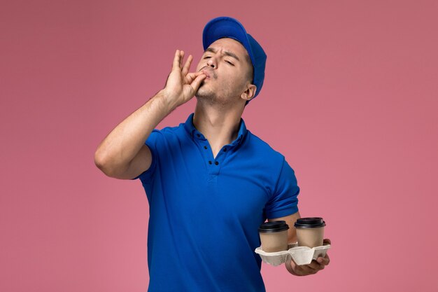 male courier in blue uniform holding coffee cups showing tasty sign on pink, uniform service delivery