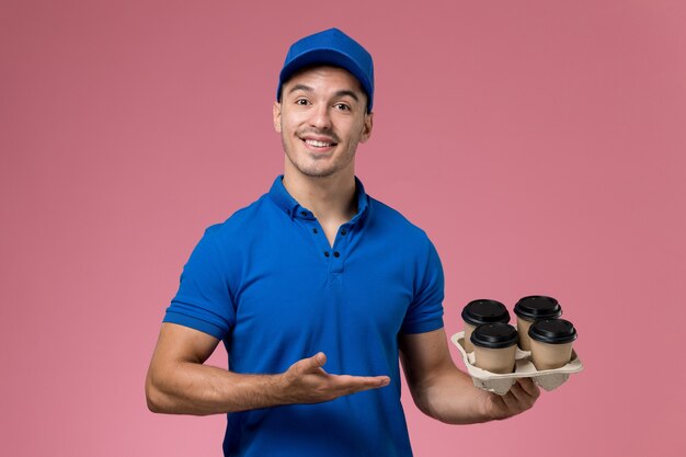 male courier in blue uniform holding coffee cups with a smile on pink, uniform service delivery worker