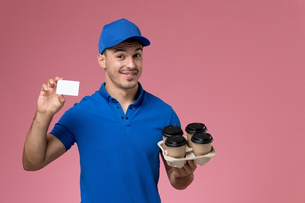 male courier in blue uniform holding delivery coffee cups and card on pink, uniform job worker service delivery