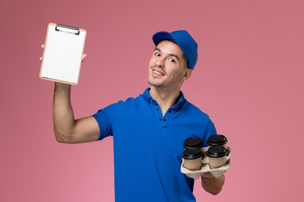 male courier in blue uniform holding delivery coffee cups and notepad on pink, uniform service delivery