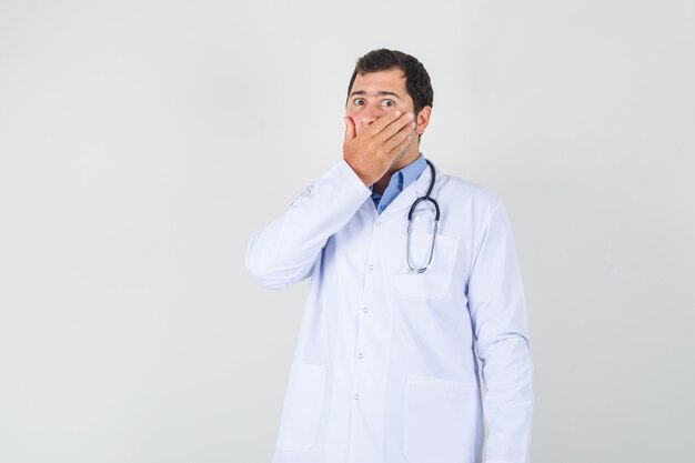 Male doctor covering mouth with hand in white coat and looking surprised. front view.