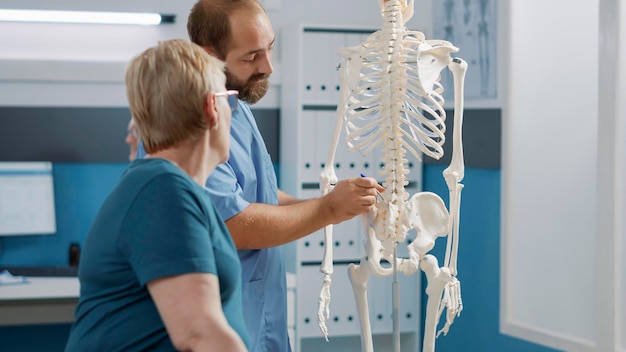 Free Photo male doctor pointing at human skeleton to show spinal cord and explain mechanical disorders in rehabilitation cabinet. specialist explaining back bones system for physical therapy.