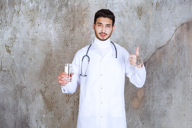 Male doctor with stethoscope holding a glass of pure water and showing positive hand sign.