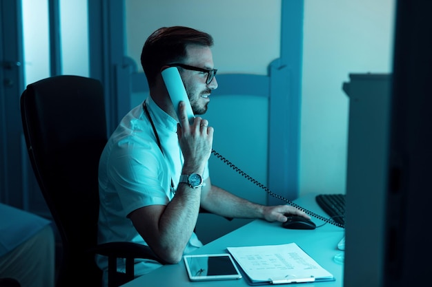 Male doctor working on a computer and communicating over the phone in the hospital