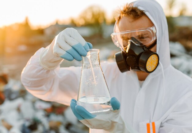 Male ecologist in radiation suit, gas mask. Holding test tube with liquid while studying trash dump.