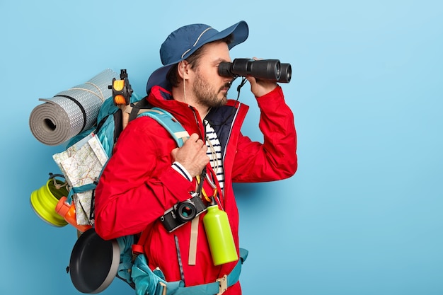 Free photo male explorer dressed in casual wear, keeps binoculars near eyes, wears hat and jacket, hikes in mountains