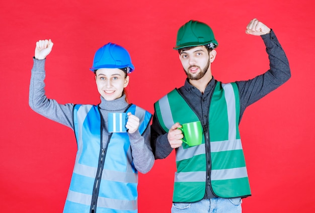 Free Photo male and female engineers with helmet holding blue and green mugs and showing satisfaction sign.