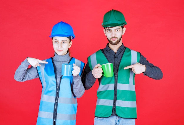 Free photo male and female engineers with helmet holding blue and green mugs.