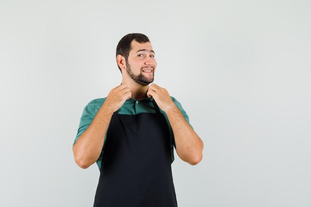 Male gardener holding his collar in t-shirt, apron and looking cheerful , front view.