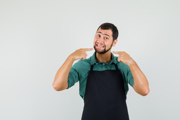 Male gardener in t-shirt, apron pointing at his teeth , front view.