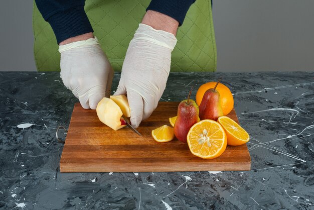 Male hand cutting apple with knife on top of wooden board on table.