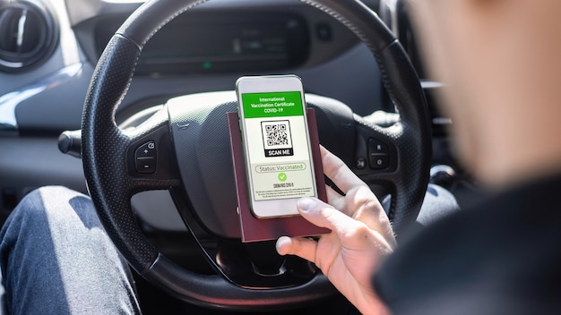 Male hand holding passport and smartphone with International Vaccination Certificate COVID-19 QR code in a car