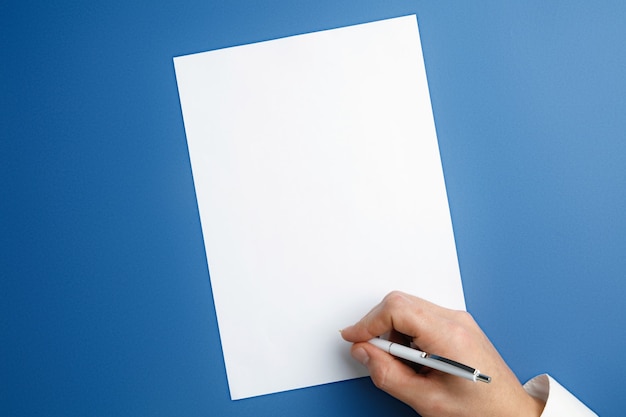 Free photo male hand holding pen and writing on empty sheet on blue wall for text or design. blank templates for contact, advertising or use in business. finance, office, purchases.  copyspace.