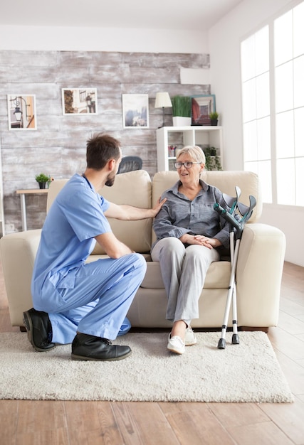 Male health visitor in a nursing home talking with retired old woman while sitting on couch. Old woman with crutches.