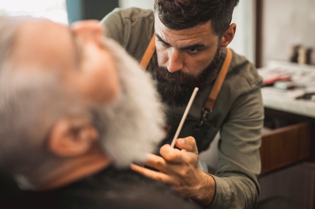 Male hipster hairdresser working with beard of senior client