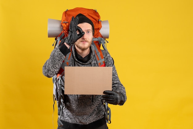 male hitchhiker with leather gloves and backpack holding blank cardboard gesturing ok sign in front of his eye