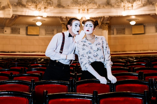 Male mime whispering in female mime's ear standing among chair