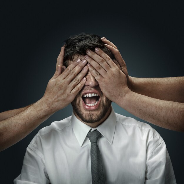 A male model surrounded by hands like his own thoughts on dark wall. A young man doubts, can't choose the right decision. Concept of mental problems, troubles in work, freedom of speech.