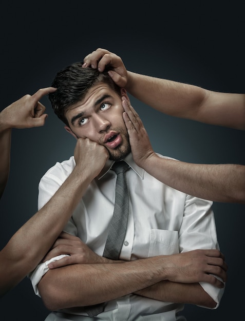 A male model surrounded by hands like his own thoughts on dark wall. A young man doubts, can't choose the right decision. Concept of mental problems, troubles in work, indecision.