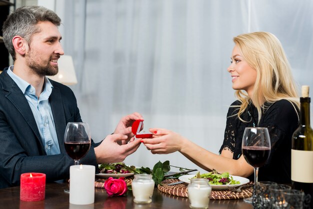 Male presenting gift box with ring to blond female at table with dishes and bloom