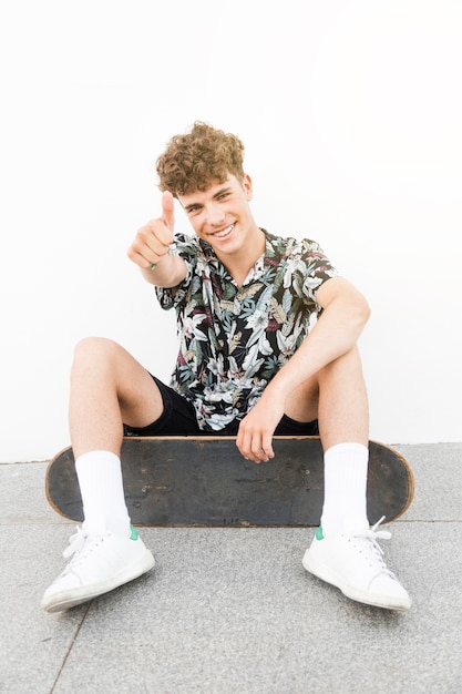 Free photo male smiling skateboarder showing thumbup sign against white wall