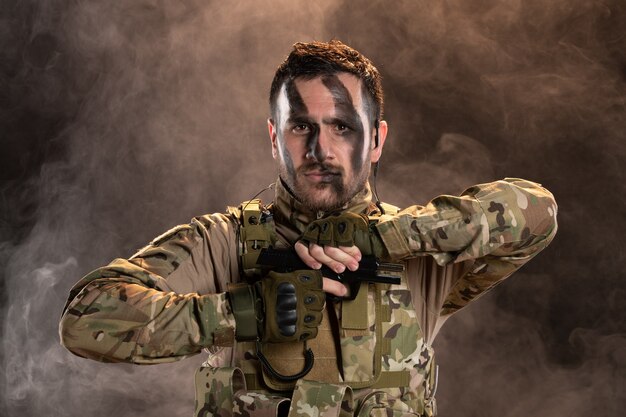 Male soldier in camouflage reloading gun on smoky dark wall