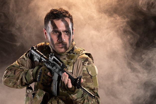 Free photo male soldier in camouflage with machine gun on a dark smoky wall