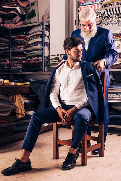 Male tailor trying the blue coat on his customer sitting over wooden chair in the shop