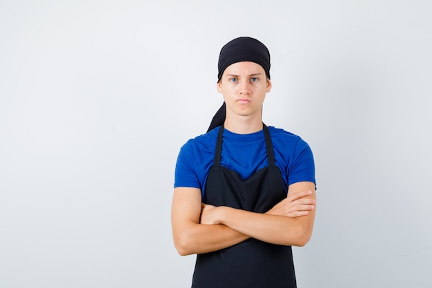 Male teen cook in t-shirt, apron with hands crossed and looking displeased , front view.