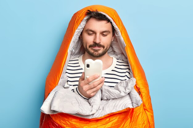 Male traveler poses in orange warm sleeping bag, spends leisure time near sea, concentrated in smartphone, finds right destination poses indoor