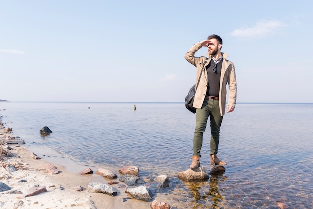 Free photo male traveler standing near the lake shielding his eyes