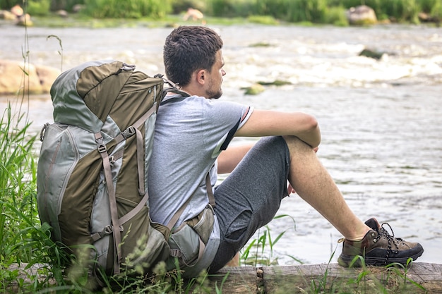 A male traveler with a large hiking backpack sits resting near the river.