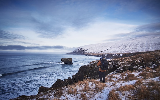 Free Photo male wearing a backpack and a jacket standing on the snowy hill while taking a picture of the sea