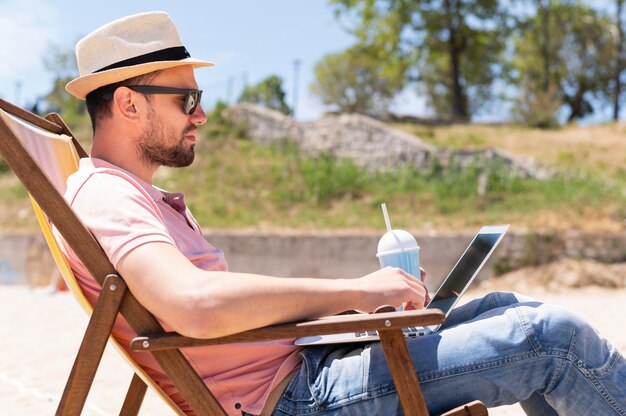 Man in beach chair working on laptop while having a drink